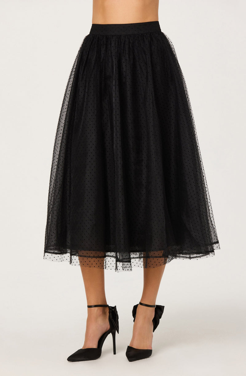 A woman is wearing a black midi-length tulle skirt with a polka dot pattern. The skirt has a fitted waistband and is made of sheer, layered tulle fabric that creates a full, flowing silhouette. She is also wearing black high-heeled shoes with ankle straps tied into bows.