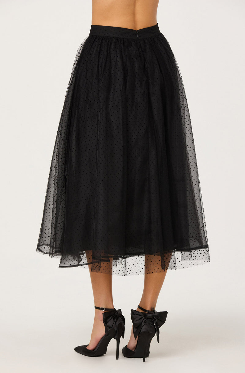 A woman is wearing a black, high-waisted, midi-length tulle skirt with a dotted pattern. The skirt is sheer with an opaque lining underneath. She is also wearing black high-heeled shoes with large bows on the ankles.