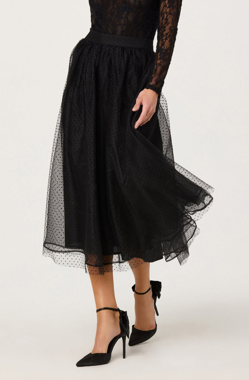 A person is wearing a black long-sleeved lace top with a floral pattern, paired with a high-waisted, black tulle skirt that falls below the knees. The skirt features a subtle dotted pattern and has a sheer overlay. They are also wearing black high-heeled shoes with ankle straps tied into bows.
