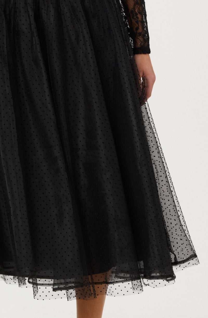 A woman is wearing a black dress with a sheer overlay featuring small polka dots. The dress has lace details on the long sleeves, and the skirt appears to be layered with tulle or similar fabric, giving it a voluminous and textured appearance.