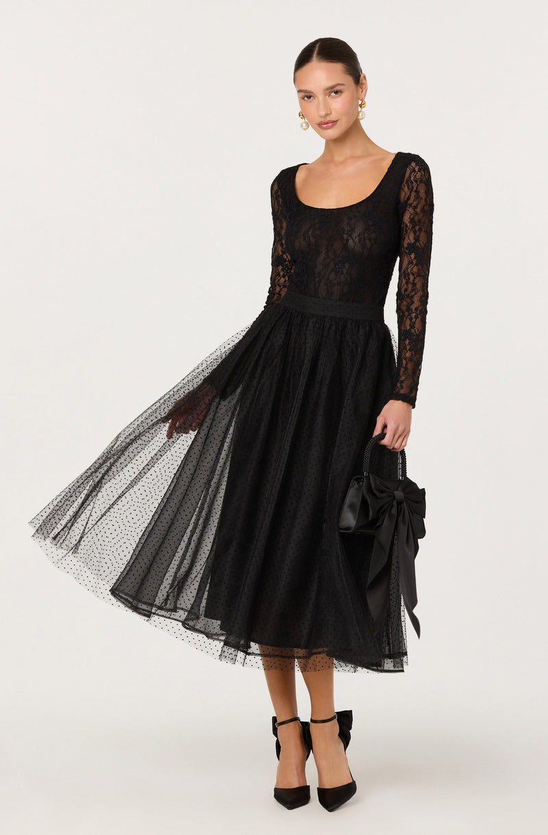 The model is wearing a long-sleeved, black lace top with a scoop neckline and a black tulle midi skirt with a dotted pattern. She is holding a black handbag with a large bow detail and wearing black ankle-strap high heels.