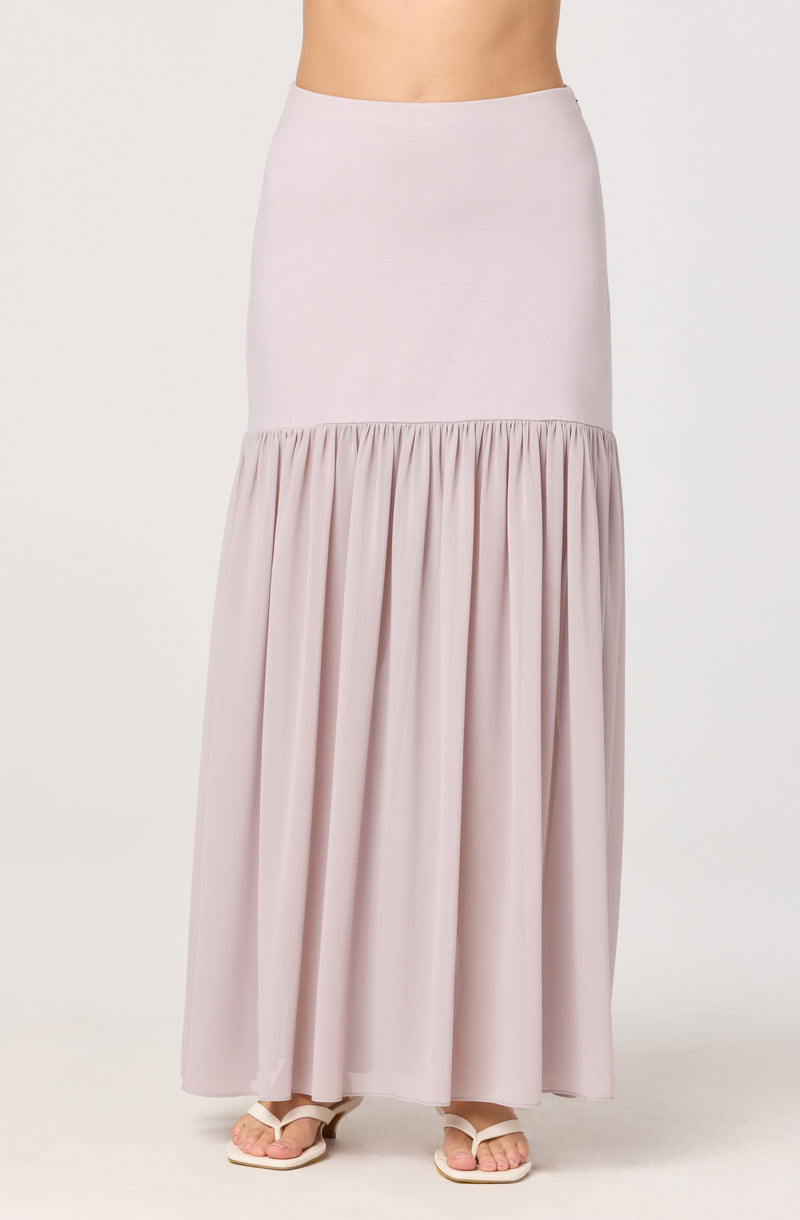 A woman is wearing a long, light pink skirt with a fitted waistband. The skirt has a smooth, solid upper portion and a gathered, flowing lower portion. She is also wearing white open-toe sandals.