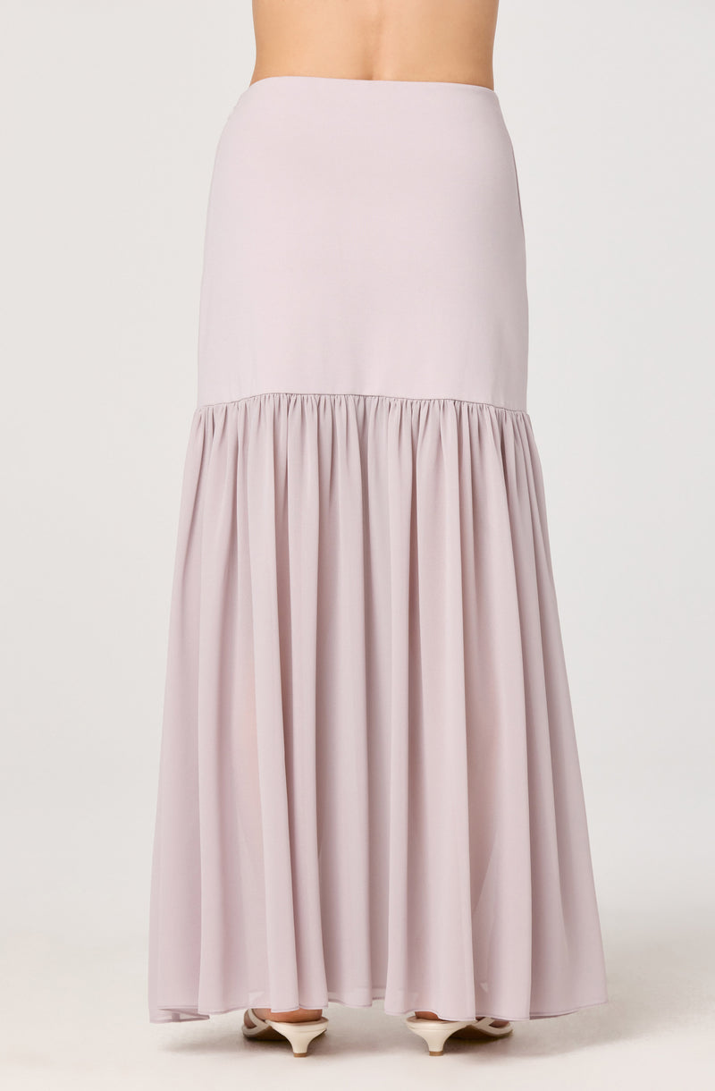 A long pastel pink skirt with a fitted waistband and a gathered, flowing lower portion that extends to the floor. The skirt has a smooth, plain upper section and a ruched, voluminous lower section with pleats. The model is wearing white shoes with a slight heel underneath the skirt.