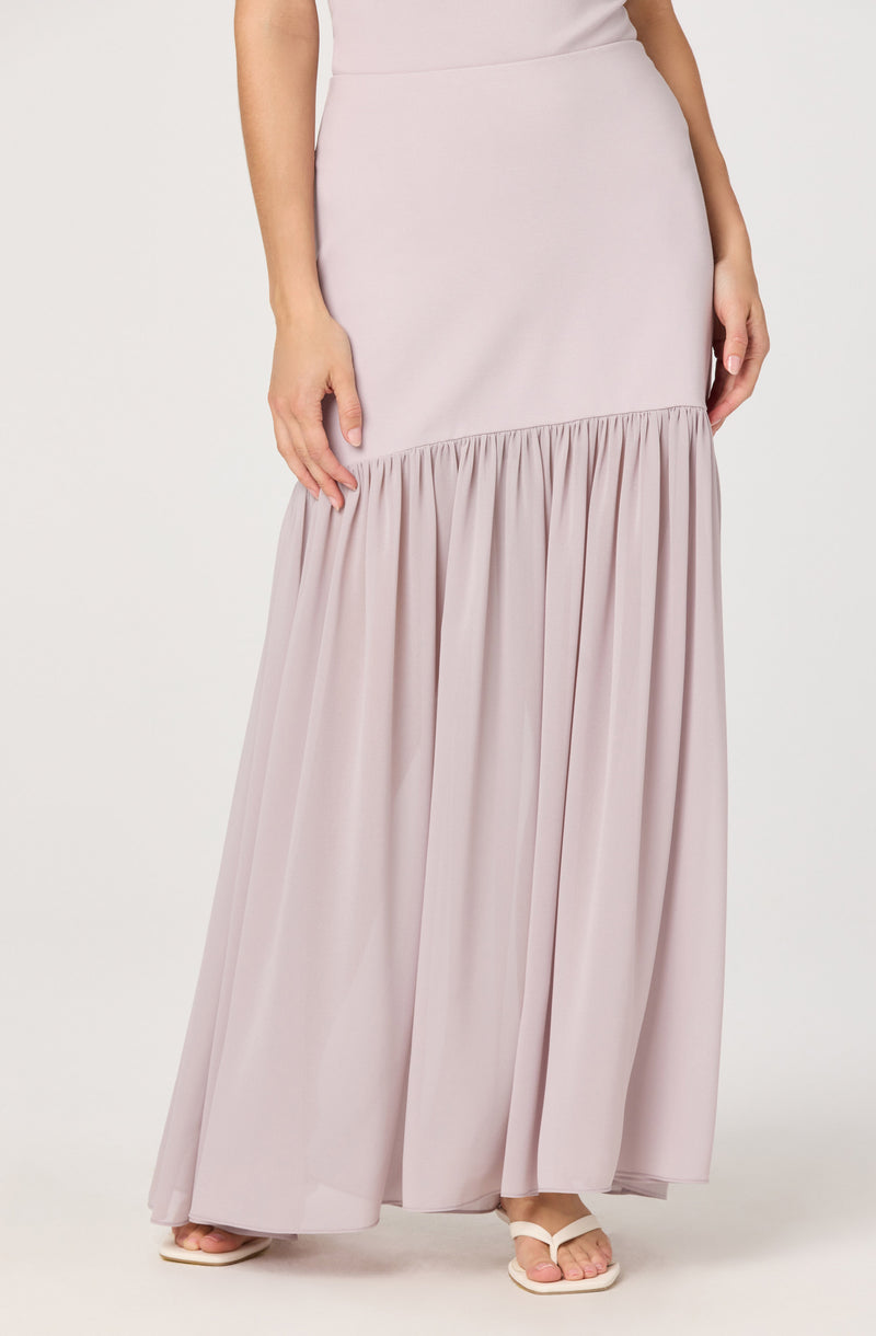 A woman is wearing a light pink, strapless maxi dress with a fitted bodice and a gathered, flowing skirt. The skirt features a ruffled seam around the midsection, creating a layered look. She is also wearing white open-toed sandals.