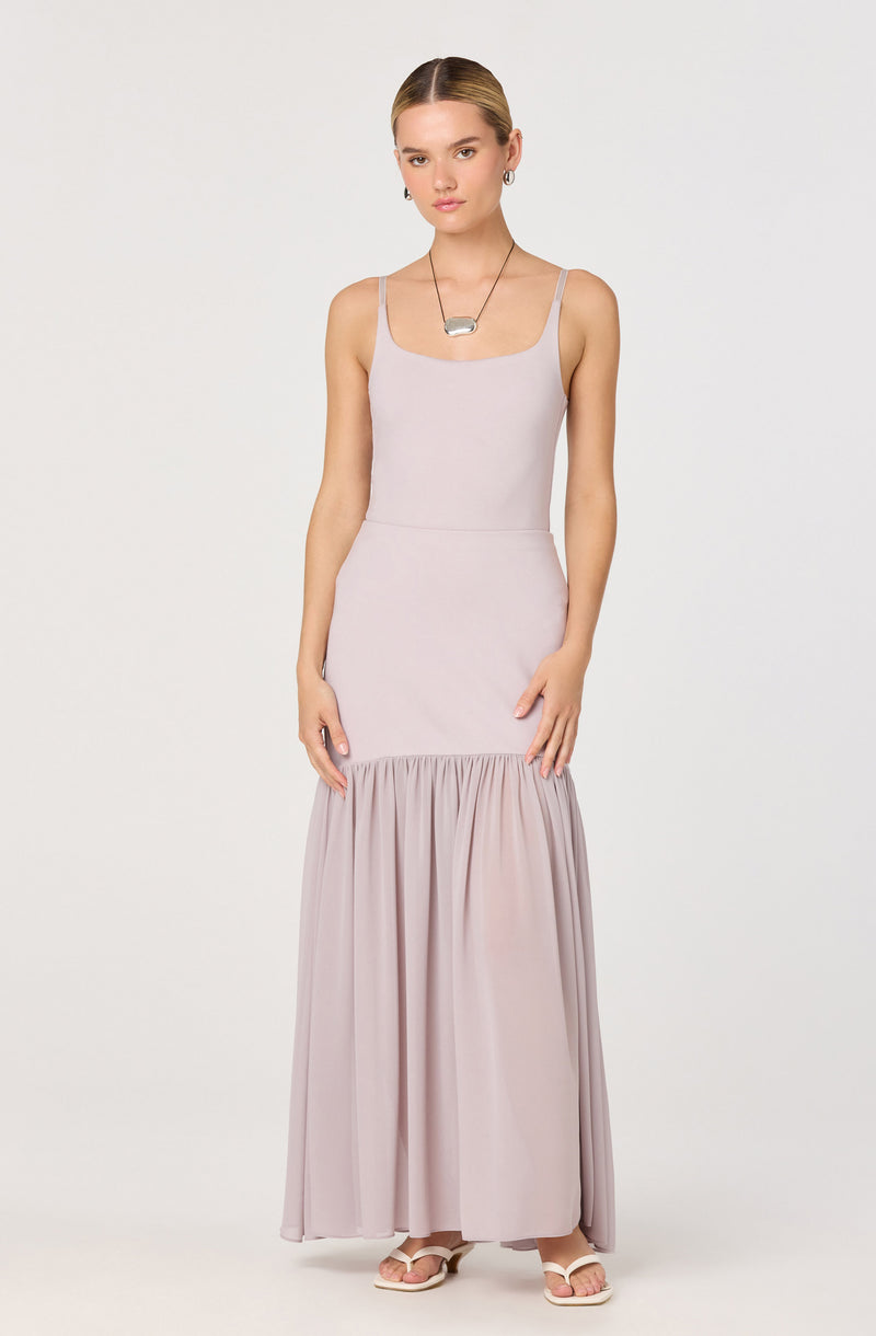 A woman is wearing a light lavender sleeveless maxi dress with thin spaghetti straps. The dress features a fitted bodice and a gathered, flowy section from the waist down. She is accessorized with a silver necklace and small earrings.