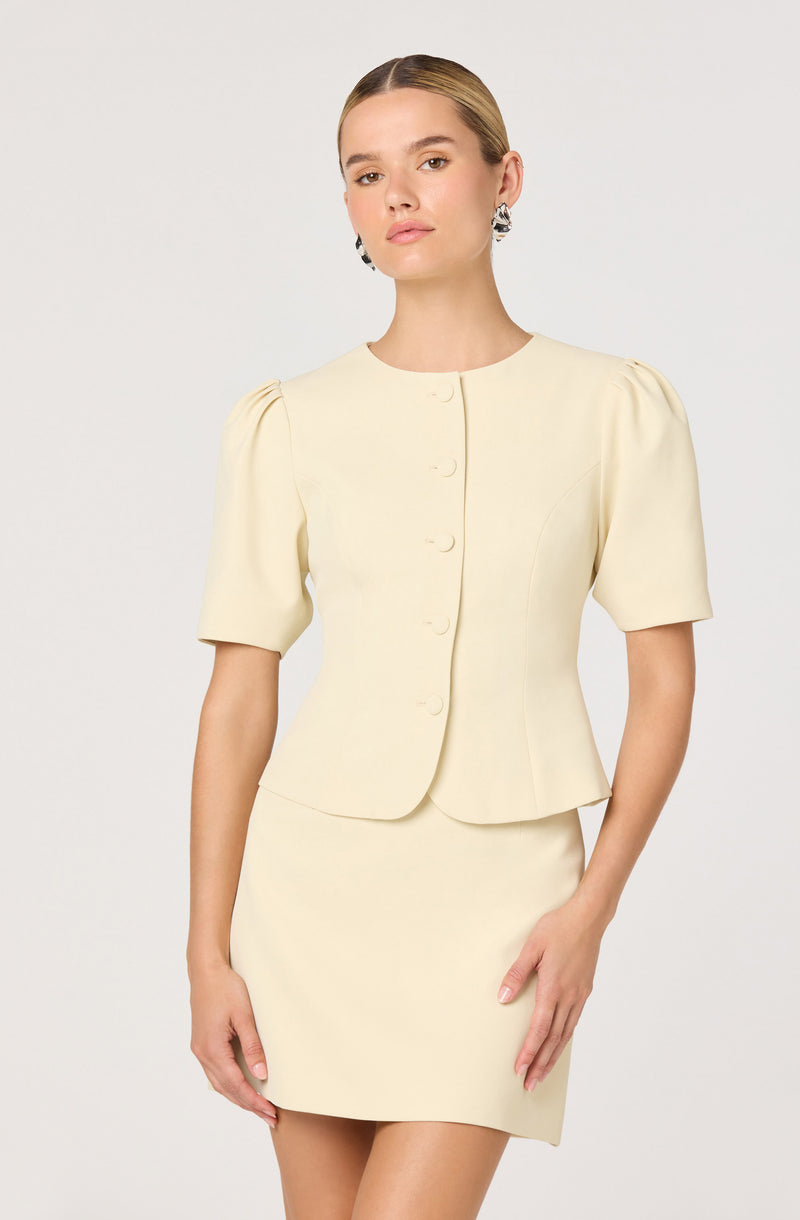 A woman is wearing a cream-colored, short-sleeved dress with a fitted waist and a matching sleeveless, button-down jacket with a rounded neckline and puffed sleeves.
