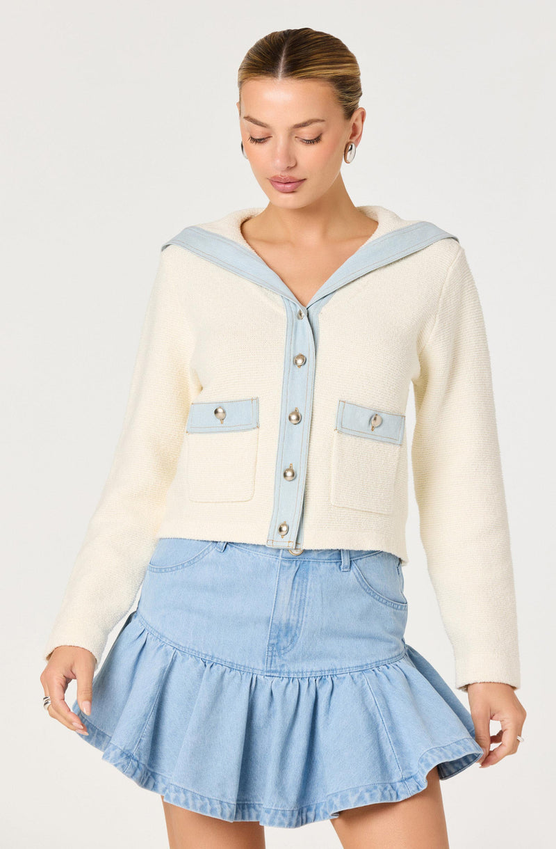 Woman in a cream sweater with light blue trim and pockets, paired with a light blue ruffled denim skirt.