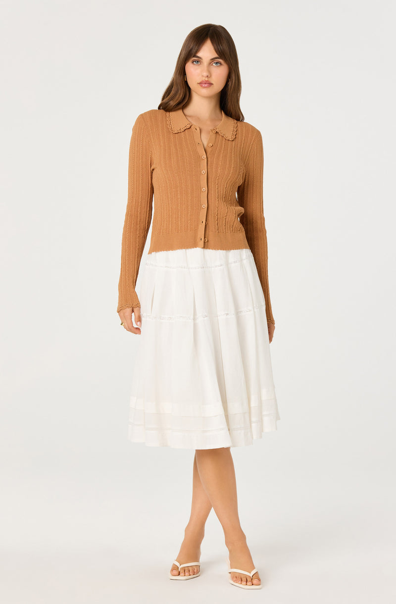 Girl wearing a light brown, button-up, long-sleeve knitted top with a scalloped collar and a white, pleated, midi skirt.