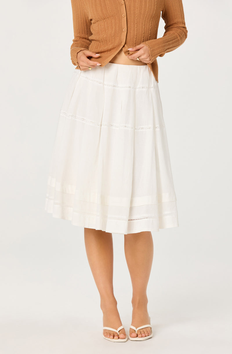 Woman wearing a brown ribbed cardigan and a white pleated midi skirt with open-toe heels.