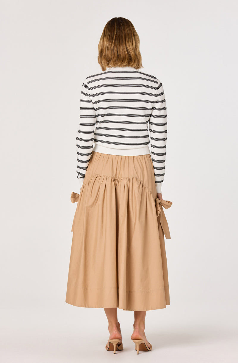 Woman wearing a white and black striped long-sleeve top and a beige, ankle-length skirt with side bows.
