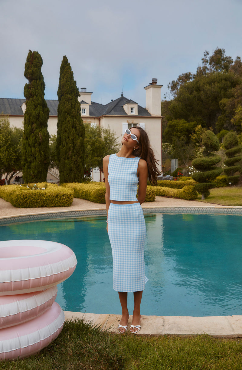 Woman in blue gingham sleeveless top and matching skirt by pool, wearing white sunglasses.