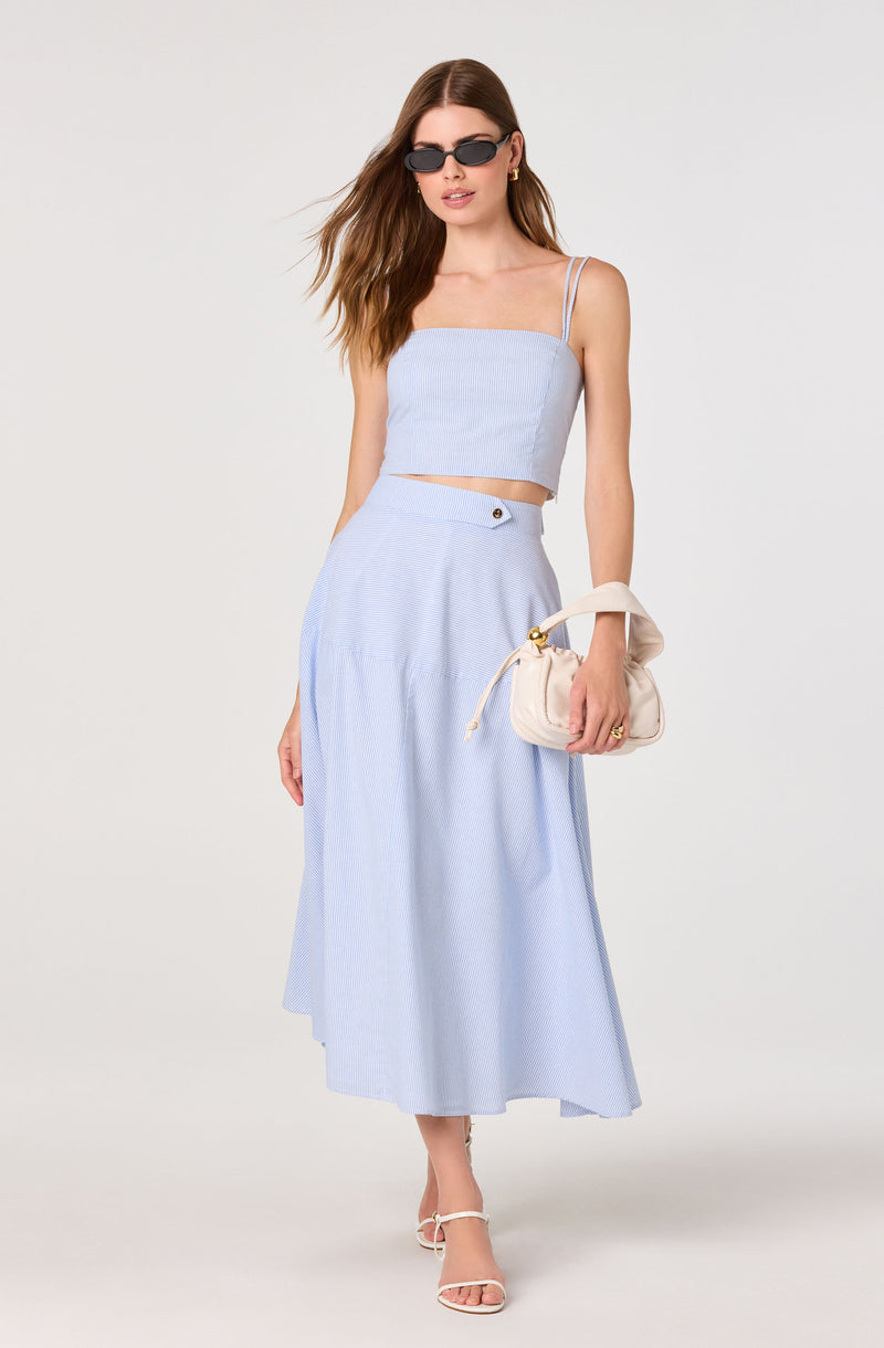 Woman in light blue two-piece outfit with spaghetti straps, paired with white heels, sunglasses, and a cream handbag.