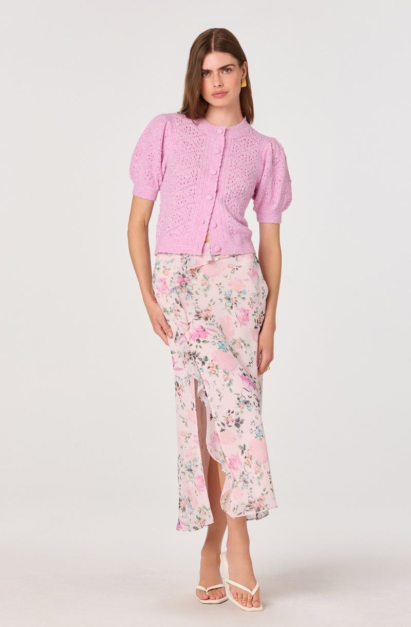 Woman in pink knitted short-sleeve cardigan with puffed sleeves and floral long skirt with front slit.
