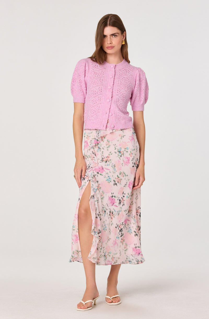 Woman in pink knit short-sleeve cardigan and floral maxi skirt with side slit.