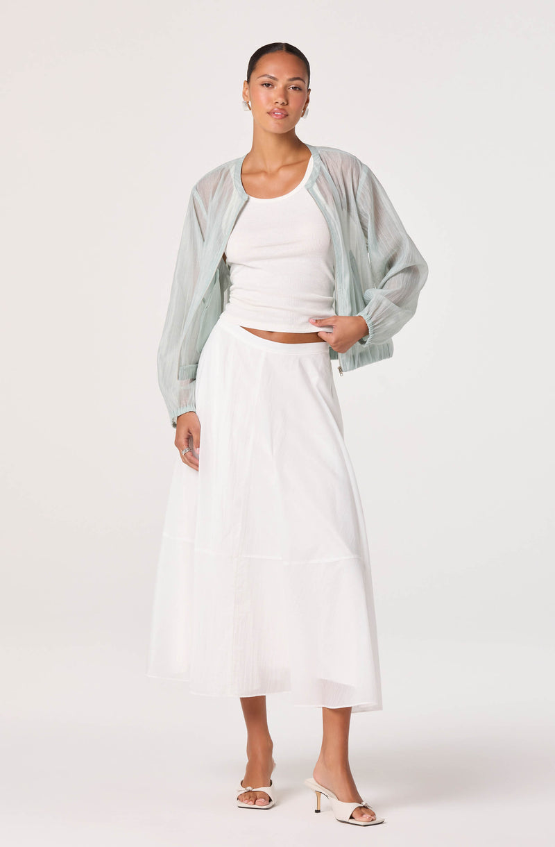 Woman wearing a sheer, light-colored bomber jacket, white crop top, and a white midi skirt.