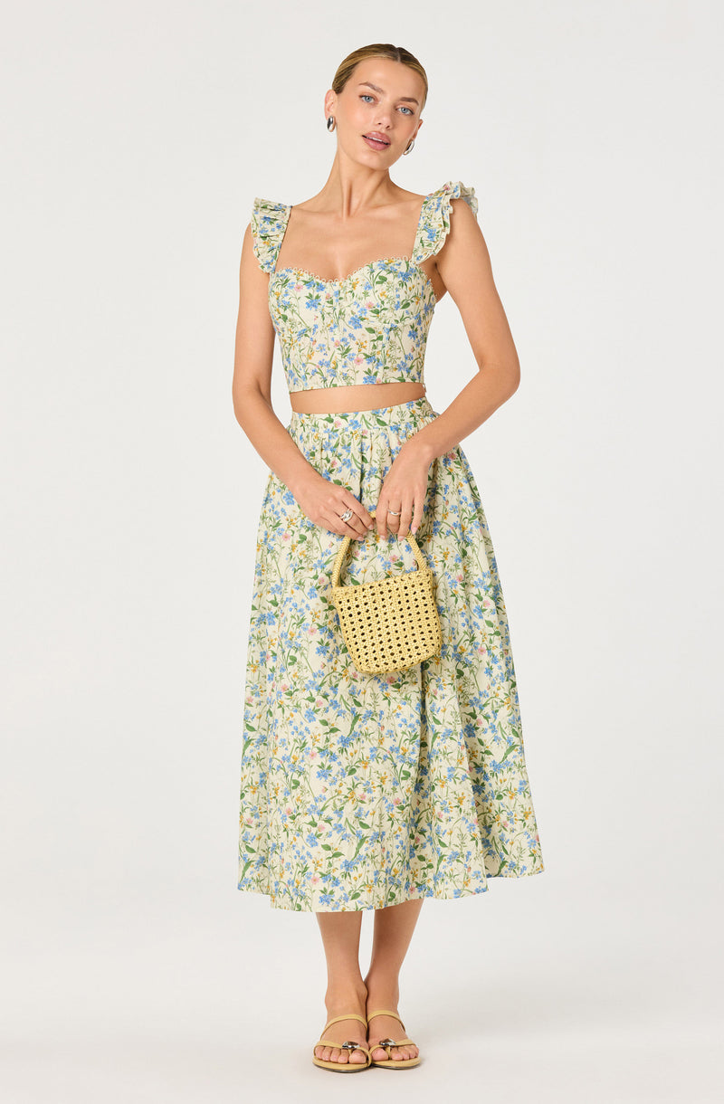 Woman in floral crop top with ruffled sleeves and matching high-waisted skirt, holding a woven yellow handbag.