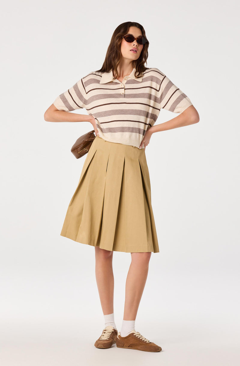 Woman wearing a beige and brown striped short-sleeve polo shirt, a beige pleated skirt, white socks, and brown sneakers.