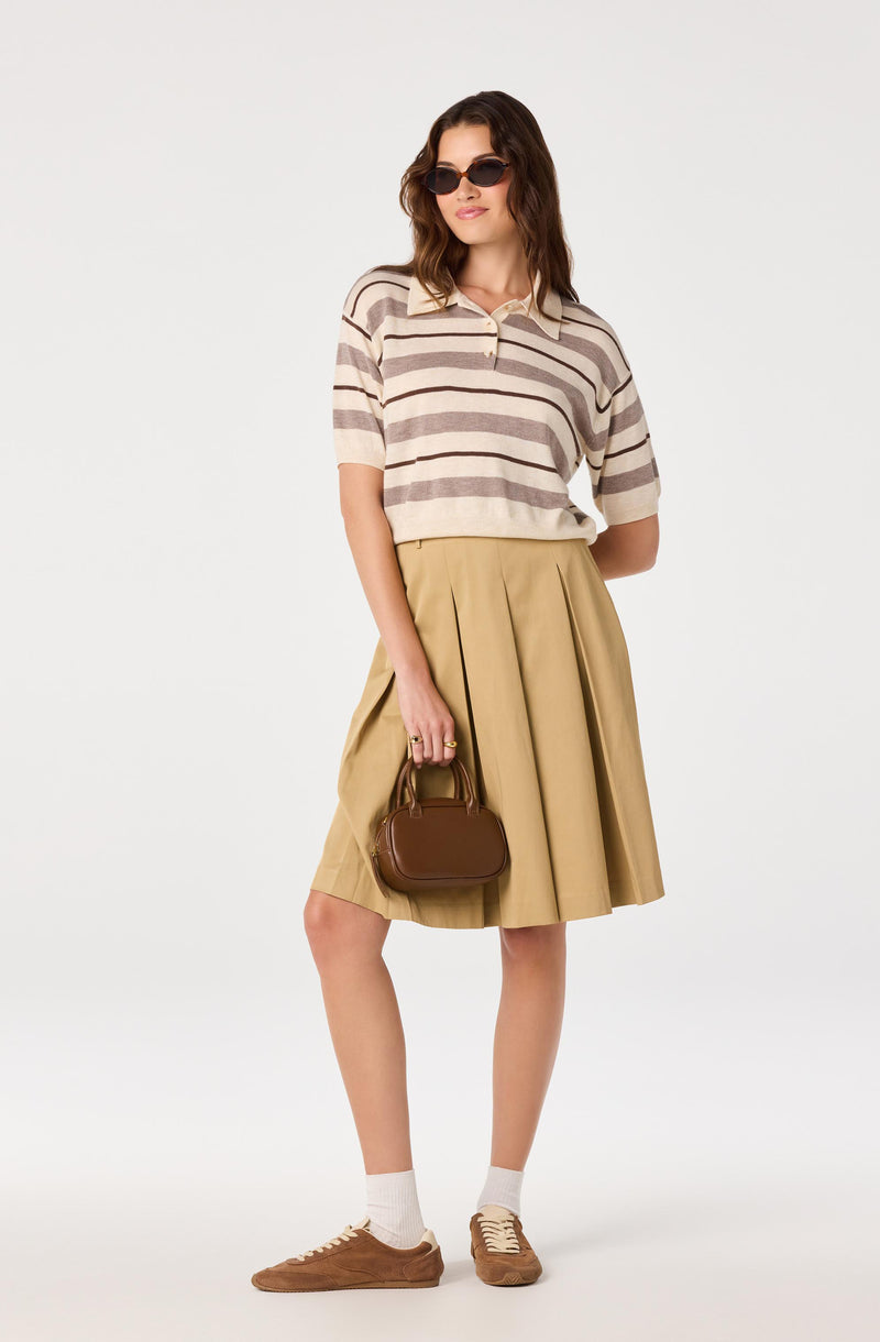 Woman wearing a beige and brown striped short-sleeve polo shirt, a pleated beige skirt, brown sneakers, white socks, and sunglasses, holding a small brown handbag.