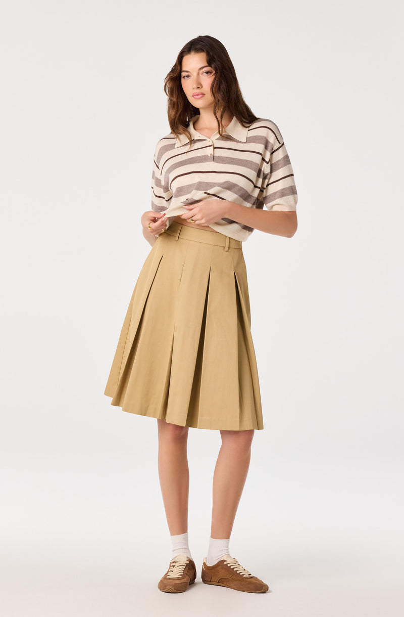 Woman wearing a beige and brown striped polo shirt, a beige pleated midi skirt, white socks, and brown sneakers.