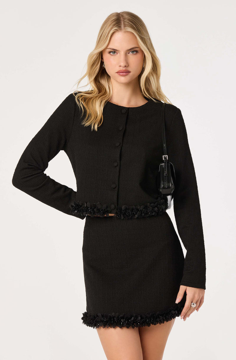 Woman wearing a black, long-sleeved, knit cardigan with button closure and floral trim, and a matching black skirt with floral trim.