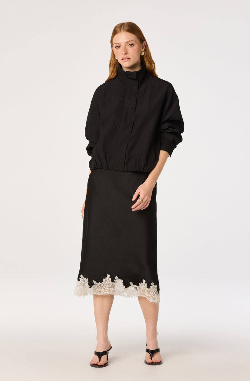 Woman wearing a black jacket with gathered sleeves and a black midi skirt with white lace trim.