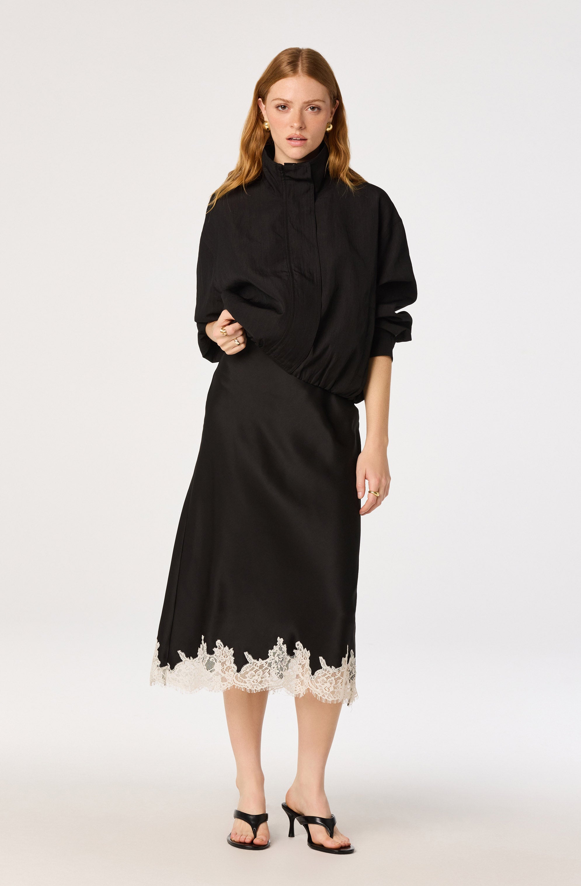 Woman in black bomber jacket and black satin skirt with lace hem, wearing black sandals.