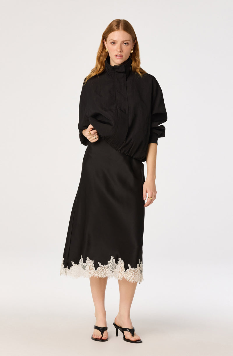 Woman in black bomber jacket and black satin skirt with lace hem, wearing black sandals.