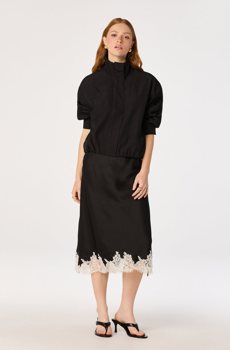 Woman wearing a black zip-up jacket with puffed sleeves and a black skirt with white lace hem.