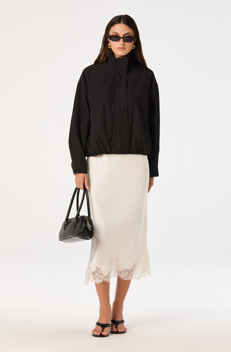 Woman in black jacket, white lace hem skirt, black sandals, holding black handbag, wearing sunglasses.