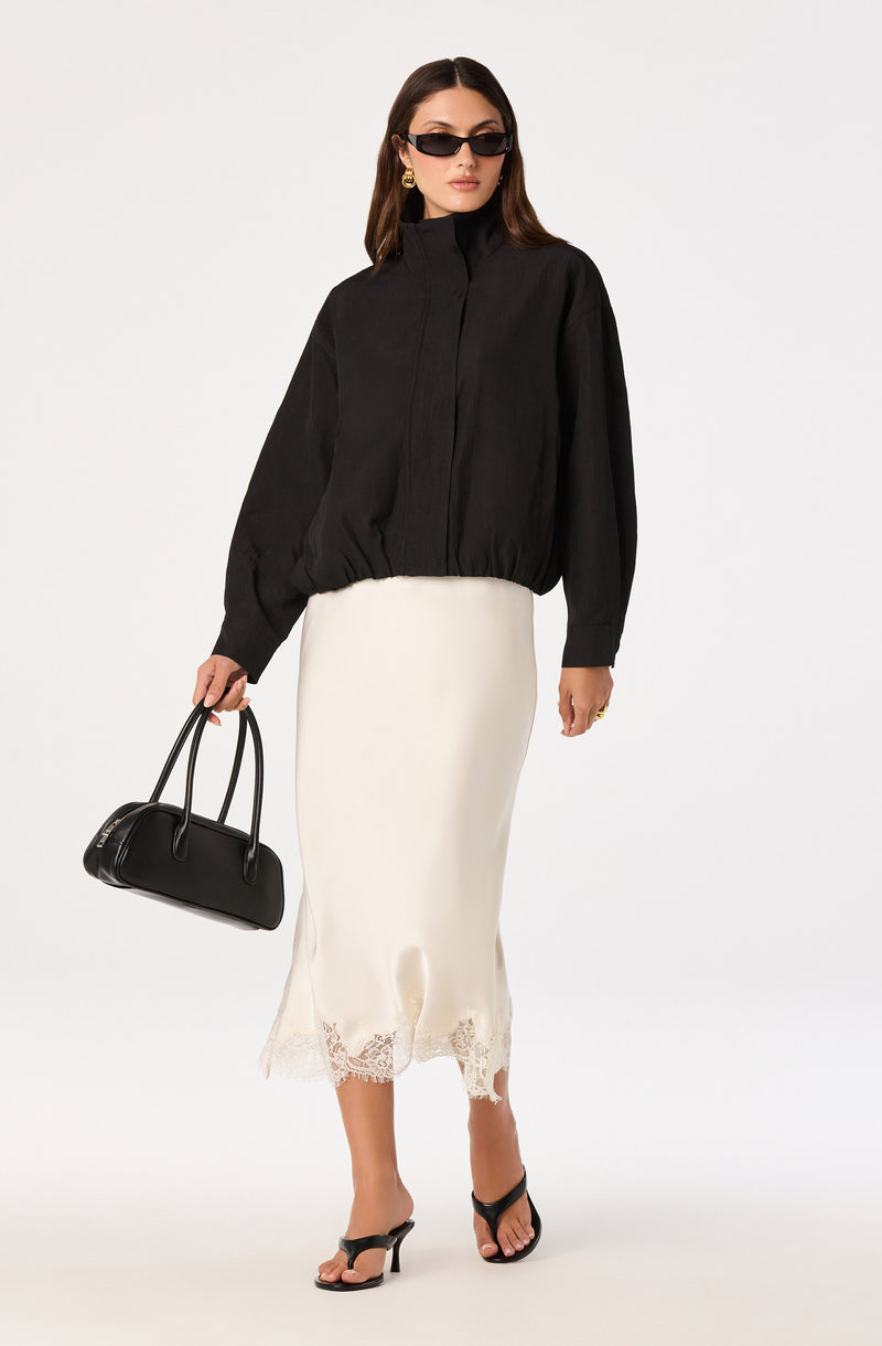 Woman wearing a black oversized jacket, white satin skirt with lace hem, black sandals, and black sunglasses.