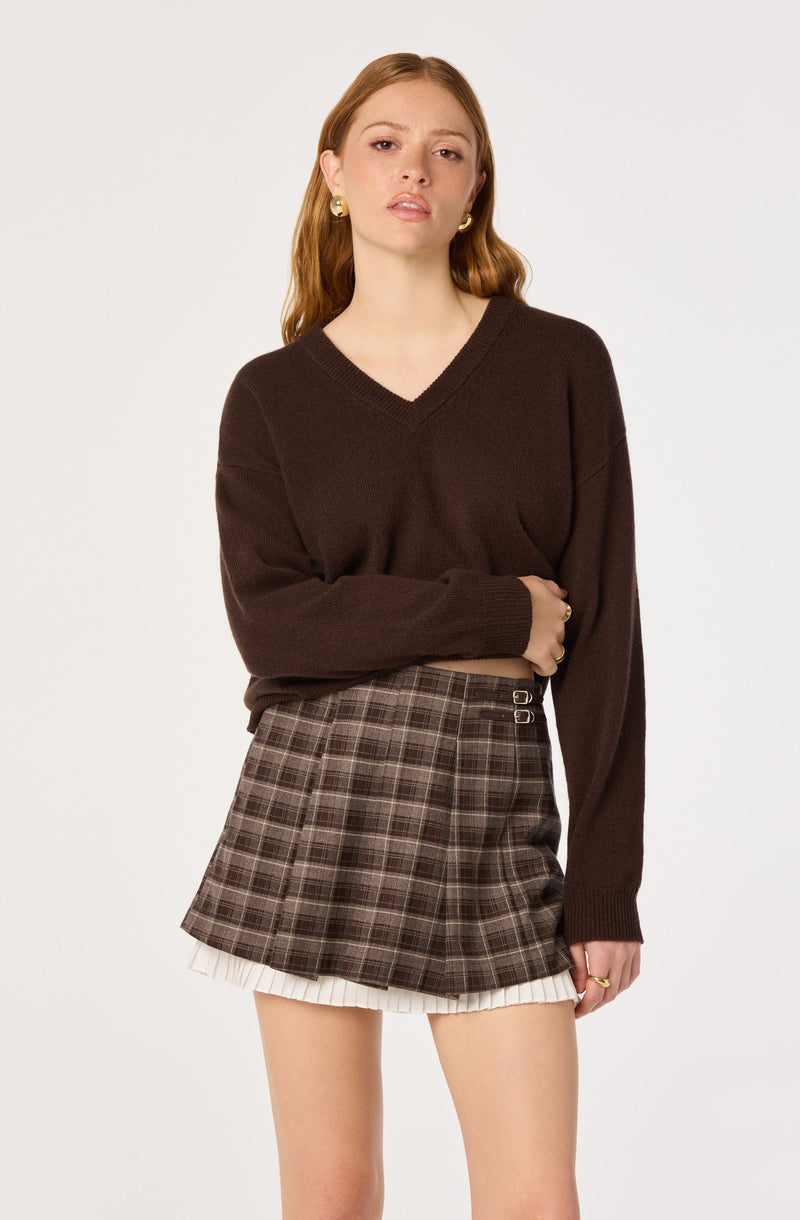 Woman wearing a brown V-neck sweater and plaid skirt with a layered white pleated underskirt.