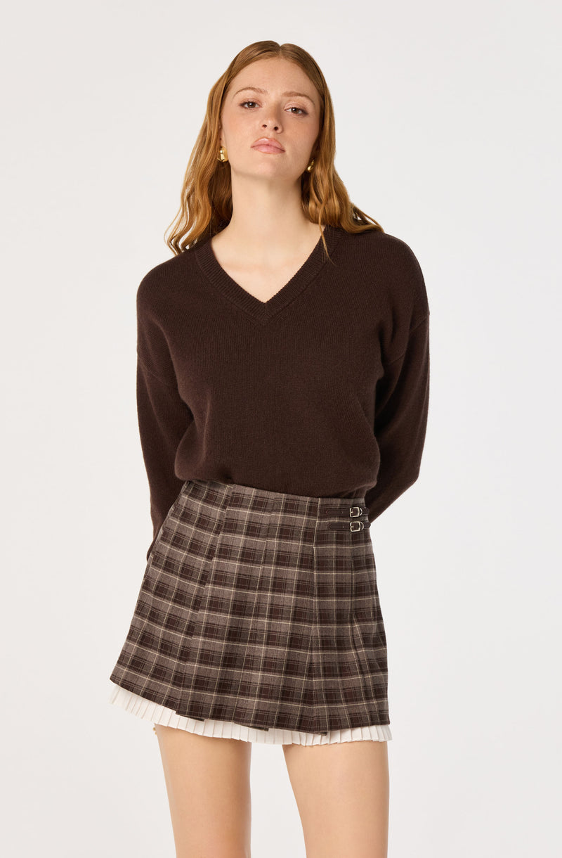 Woman wearing a dark brown V-neck sweater and a plaid skirt with pleats and buckle details.