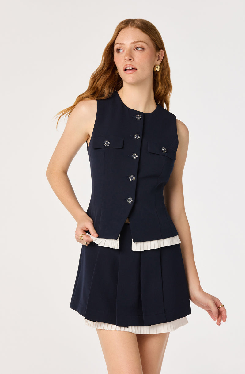 Model wearing a navy sleeveless vest with decorative buttons and matching pleated skirt with white trim.