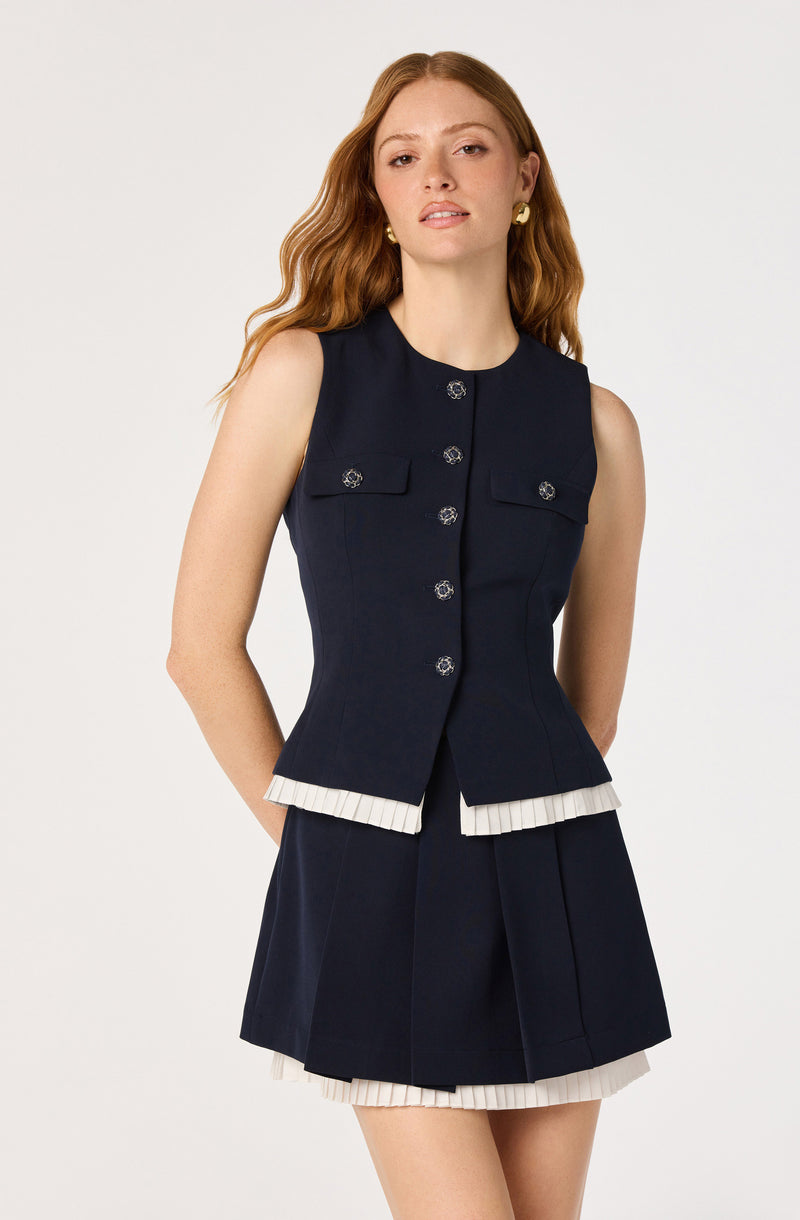 Woman wearing a sleeveless navy blue vest with silver buttons and matching navy blue skirt with white pleated trim.