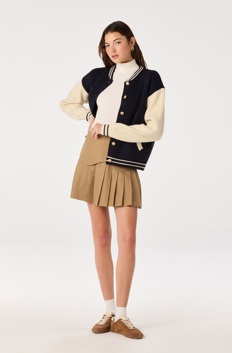 Girl wearing a navy and cream varsity jacket, white turtleneck, beige pleated skirt, white socks, and tan sneakers.
