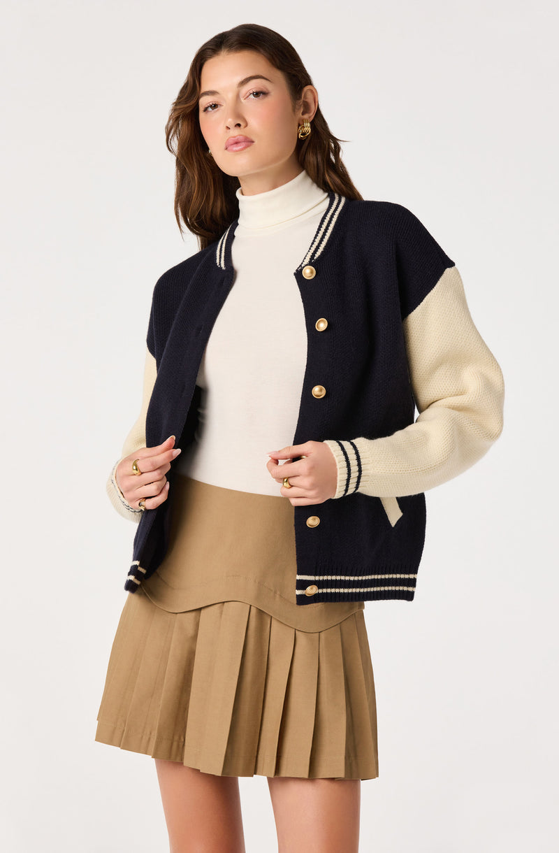 Woman wearing a cream turtleneck, navy and cream varsity jacket, and a tan pleated skirt.