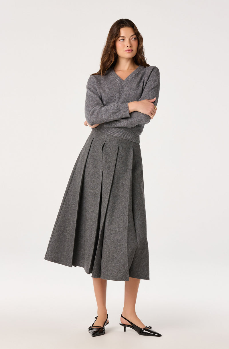 Girl in a gray V-neck sweater and high-waisted pleated gray wide-leg skirt, black shoes.