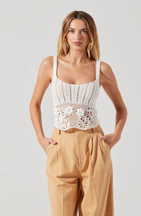 A woman is wearing a white sleeveless crochet top with thick straps and intricate lace and floral patterns at the bottom. She pairs the top with high-waisted, loose-fitting beige trousers with pleats.