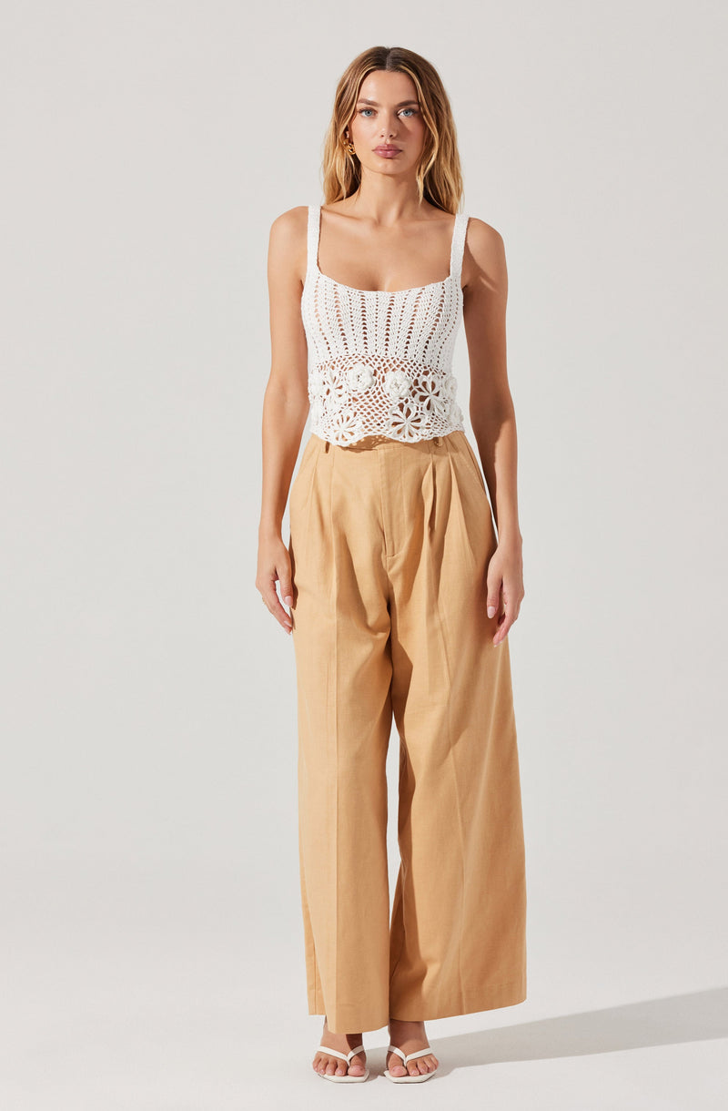 A woman is wearing a white crochet tank top with wide straps, featuring a floral pattern at the bottom edge. She pairs it with high-waisted, wide-leg beige trousers. She is also wearing white open-toe sandals.