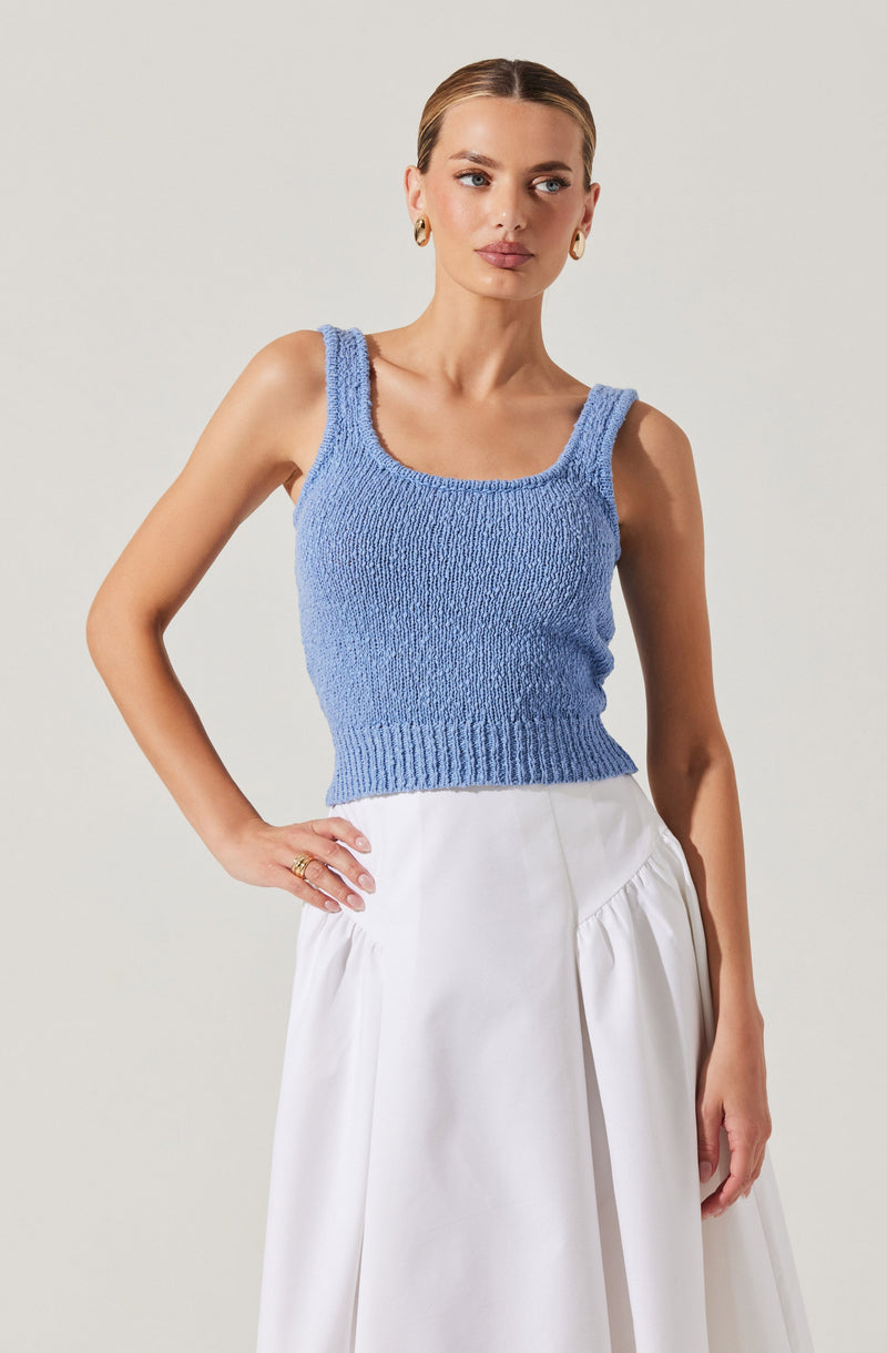 A woman is wearing a sleeveless, light blue, knitted crop top with wide straps and a ribbed hem. She pairs it with a high-waisted, white skirt that has a gathered, voluminous design. She is accessorized with gold hoop earrings and rings.