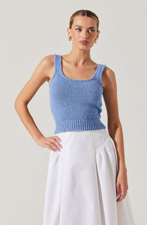 A woman is wearing a sleeveless, light blue, knitted crop top with wide straps and a ribbed hem. She pairs it with a high-waisted, white skirt that has a gathered, voluminous design. She is accessorized with gold hoop earrings and rings.
