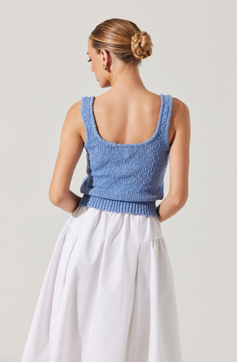 A woman is wearing a sleeveless, light blue knitted top with wide shoulder straps and a scoop neckline. She pairs it with a high-waisted, white, flowing skirt that reaches below her knees. Her hair is styled in a neat twisted bun at the back of her head.