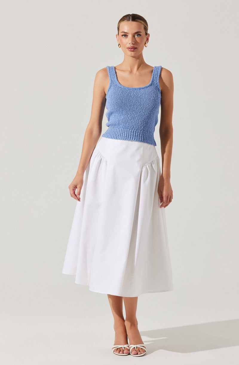 A woman is wearing a sleeveless, knitted, light blue top with wide straps and a scoop neckline. She pairs it with a white, high-waisted, ankle-length skirt that has a gathered design around the waist. She is also wearing white open-toe heels and gold hoop earrings.
