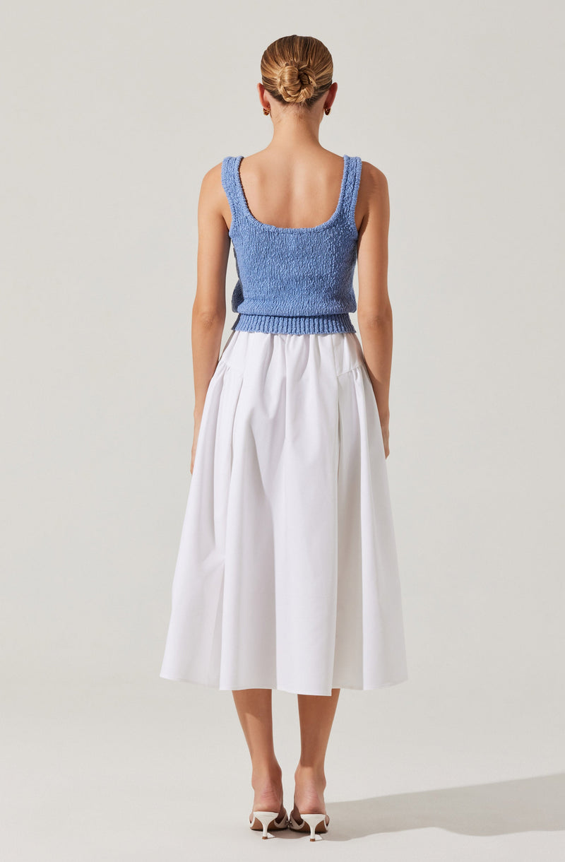 A woman is wearing a sleeveless, textured blue knit top with wide straps and a scoop neckline. She pairs it with a high-waisted, white midi-length skirt that has a full, flowing silhouette and pleats. She is also wearing light-colored, pointed-toe heels with ankle straps. Her hair is styled in an elegant updo.