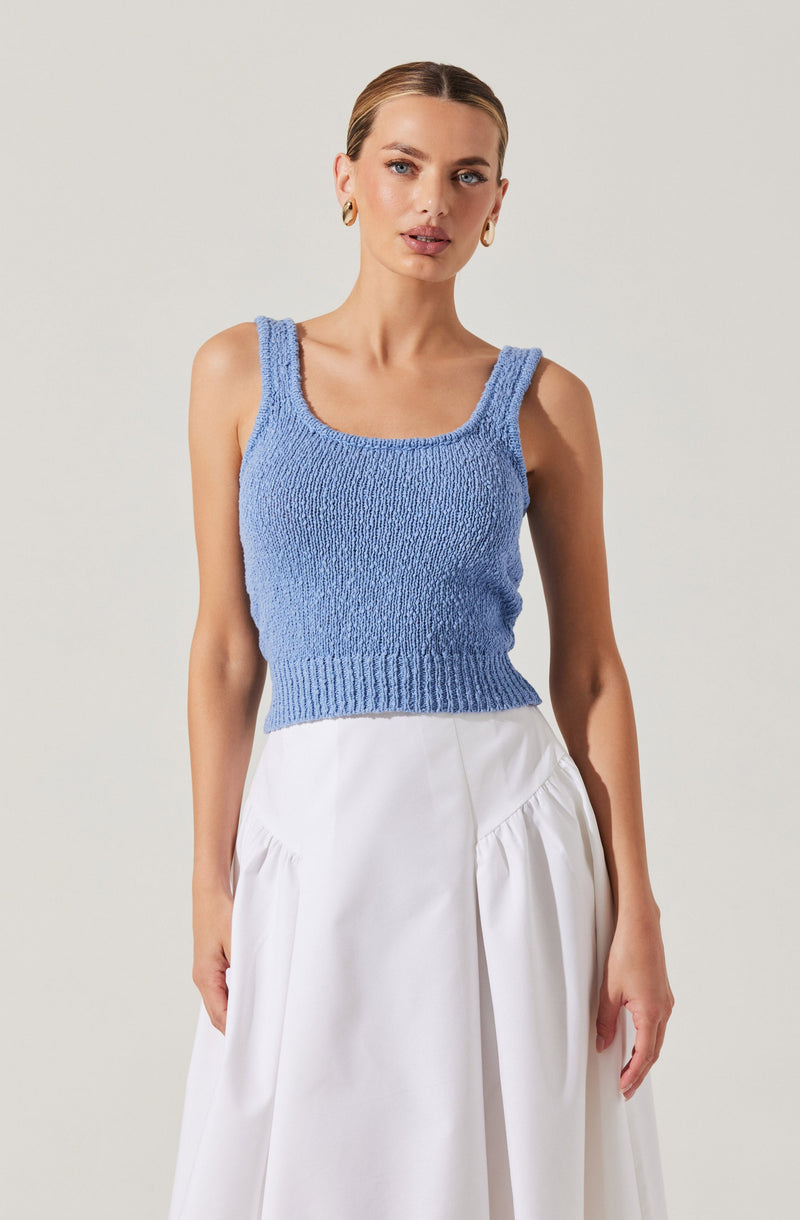 A woman is wearing a sleeveless, knitted blue tank top with wide straps and a rounded neckline. She pairs it with a high-waisted, flowy white skirt that has gathered fabric details on the sides. She is accessorized with gold hoop earrings. The background is a plain, light-colored backdrop.