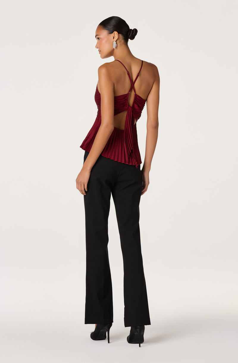 Woman wearing a burgundy pleated top with thin crisscross straps and open back, paired with black pants.