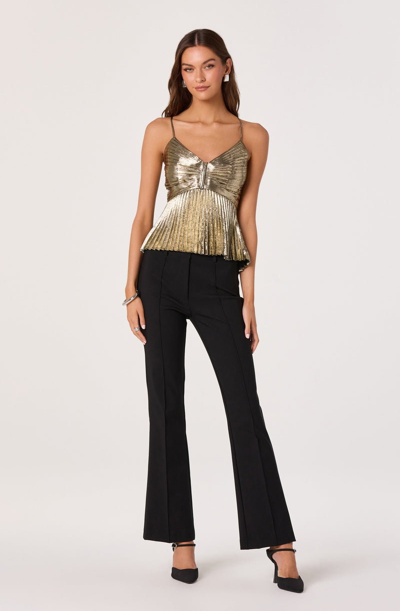 A woman is wearing a gold metallic sleeveless top with thin spaghetti straps, a ruched detail at the center, and a ruffled hemline. She pairs the top with high-waisted, black, wide-leg trousers that have front seams and a tailored fit. She completes the look with black ankle-strap heels.