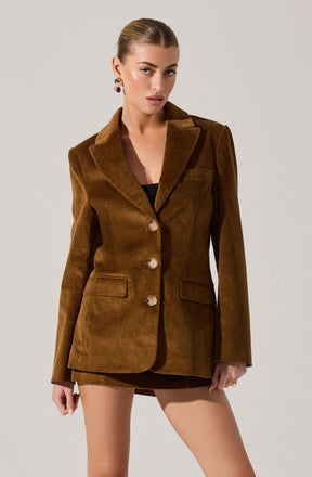 A woman is wearing a brown corduroy blazer with a notched lapel collar, long sleeves, and three large buttons down the front. The blazer features two flap pockets on the front. She is also wearing a black top underneath and has accessorized with colorful earrings.