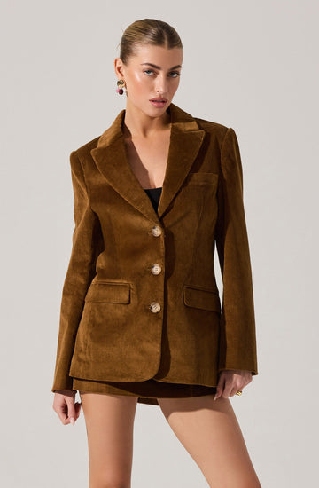 A woman is wearing a brown corduroy blazer with a notched lapel collar, long sleeves, and three large buttons down the front. The blazer features two flap pockets on the front. She is also wearing a black top underneath and has accessorized with colorful earrings.