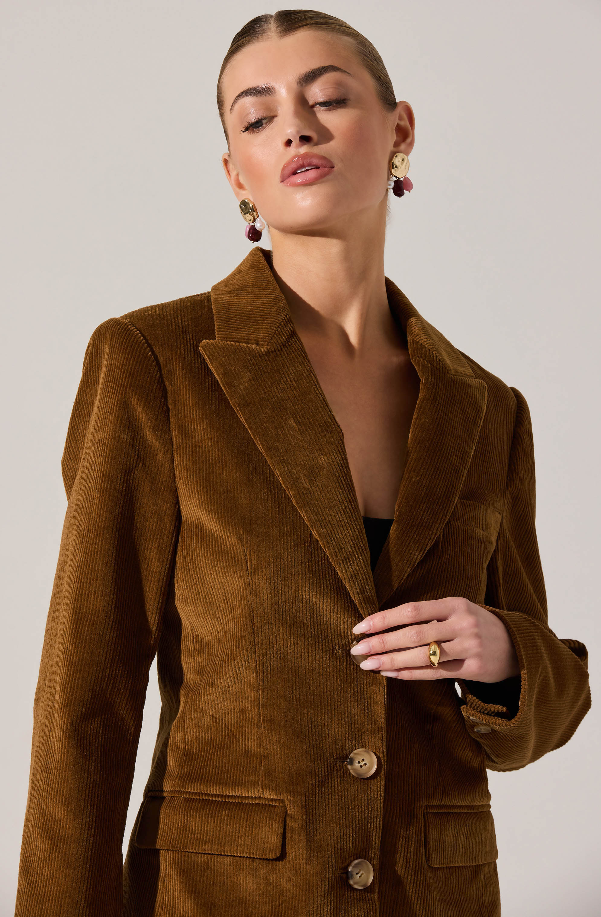 A woman is wearing a brown corduroy blazer with a notched lapel collar, two front flap pockets, and two buttons down the front. She has earrings with large, gold-colored disc and purple bead accents, and a gold ring on her right hand.